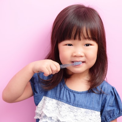 Little girl brushing her teeth and standing in front of a pink background